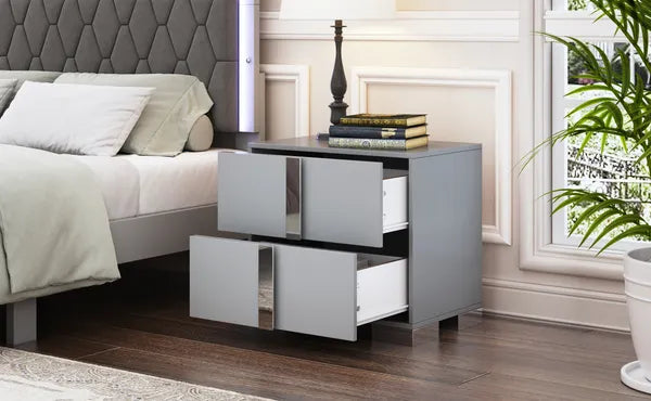 Gray mirrored nightstand with two open drawers and sleek metal feet next to a bed.