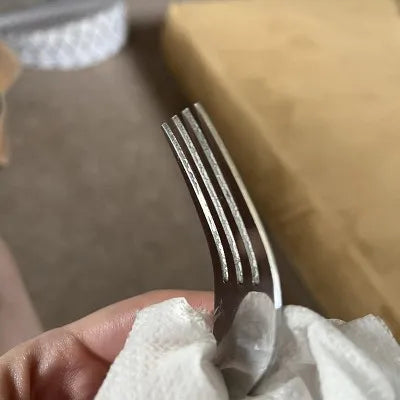 Hand holding a silver, mirror-polished stainless steel dinner fork with four tines, being wiped with a white cloth.