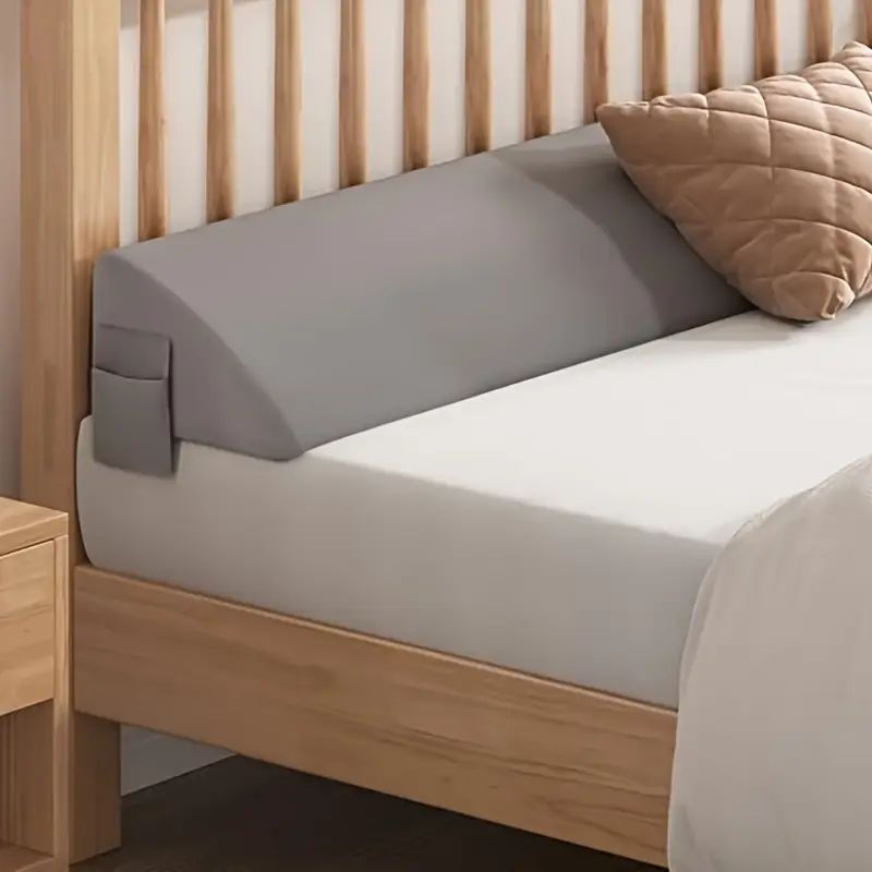 Gray ergonomic mattress gap filler with side pocket, bridging a bed and wooden headboard.