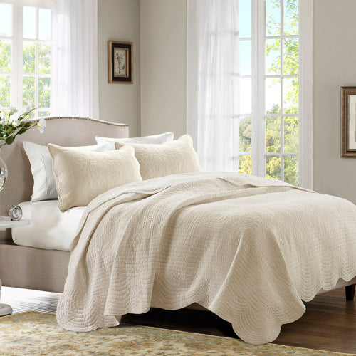 Cream reversible scalloped edge quilt set with two matching shams on a bed in a bright bedroom.
