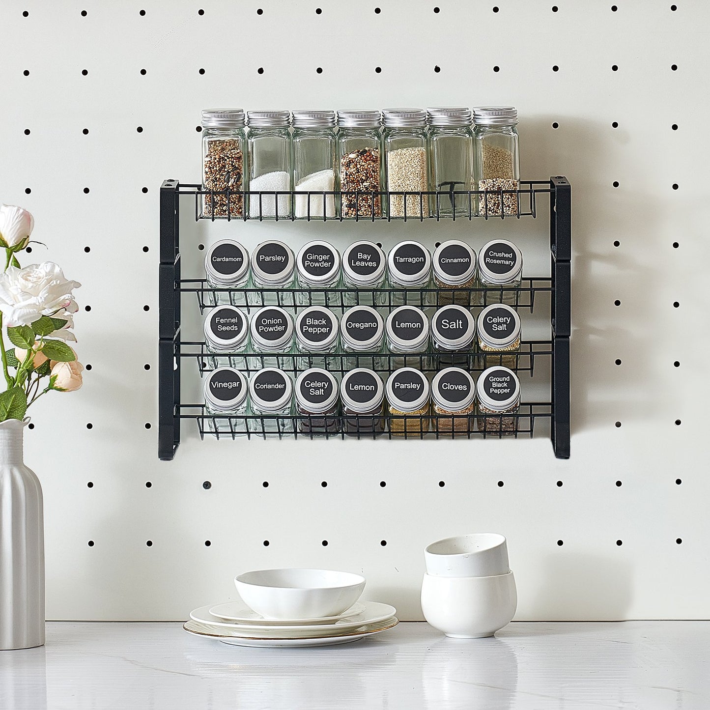 Black metal tiered spice rack with 18 labeled jars on a white pegboard, next to a white vase and dishes.